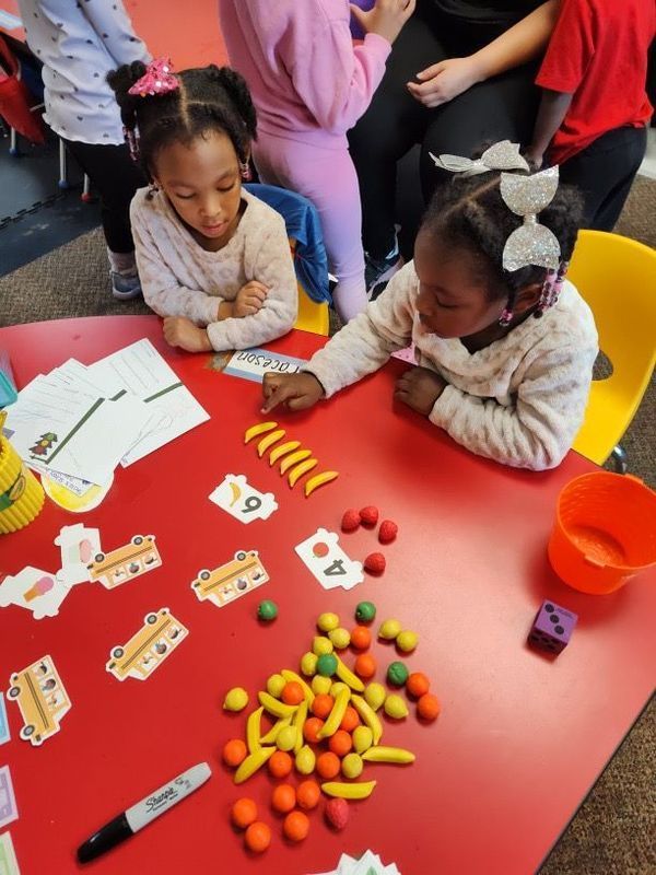 Two children at a red table with learning materials: number cards, playdough, and counters.