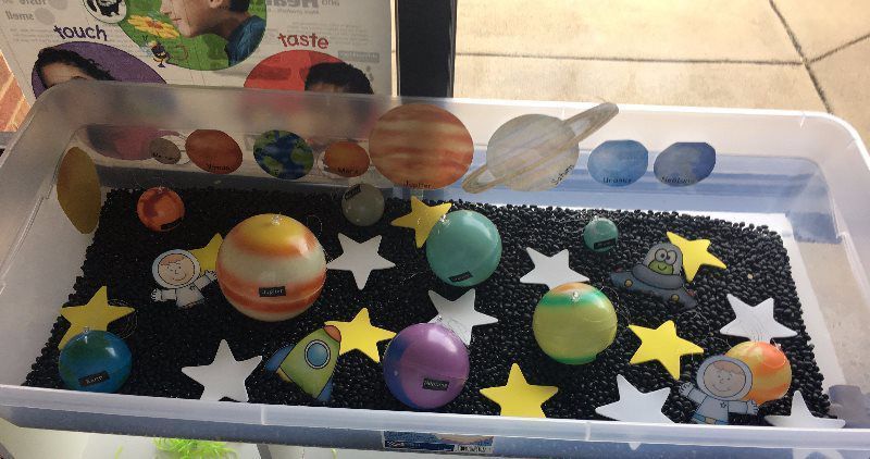 Sensory bin with planets, stars, astronaut cutouts, and black beads.