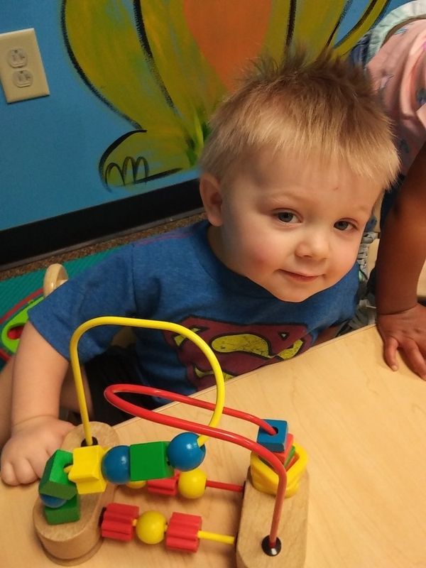 Child playing with bead maze toy, smiling, wearing blue Superman shirt.