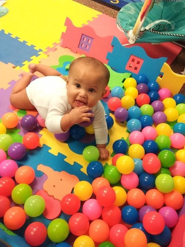 Baby in white onesie, playing in a ball pit with colorful balls on a colorful mat.