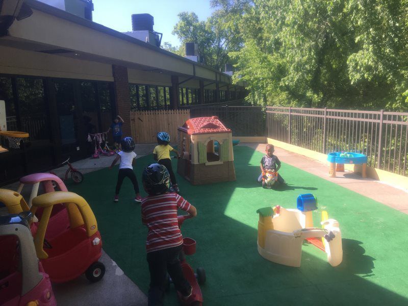 Children playing outside on a green surface with ride-on toys, a playhouse, and a fence.