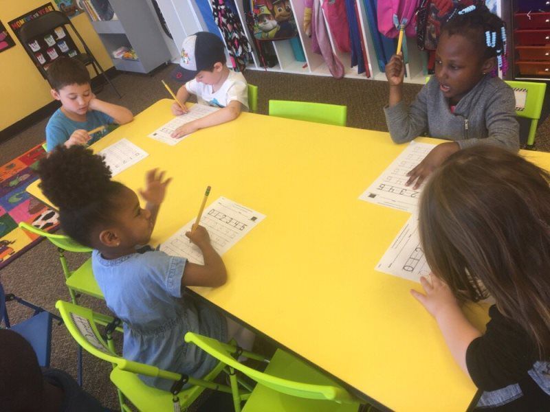 Children at yellow table writing with pencils. Bright room with colorful chairs.