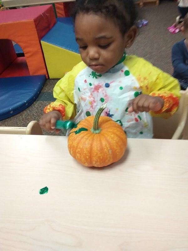 Child painting a pumpkin at a table. Wearing a paint smock, with green paint, indoors.