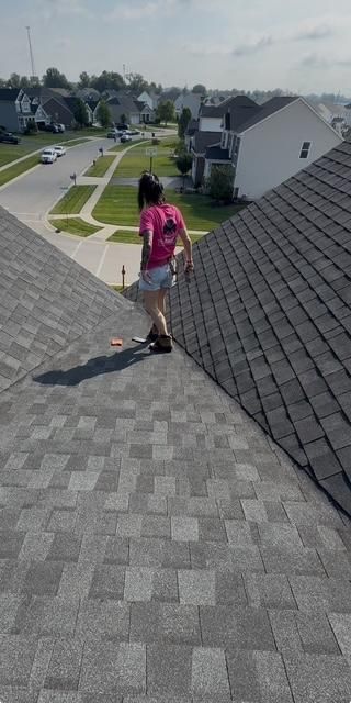 Person in pink shirt walking down a shingled roof in a suburban neighborhood.