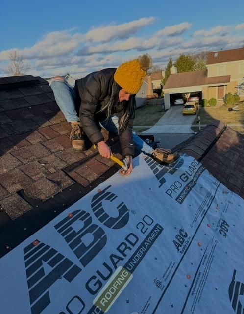 Worker installing roof underlayment on a shingled roof, wearing a yellow beanie and black jacket.