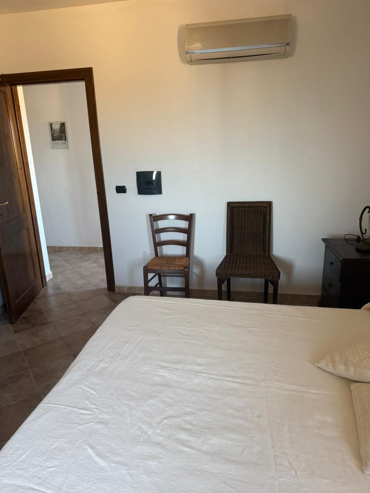 Camera da letto con letto bianco, due sedie, condizionatore a parete e porta che conduce alla stanza adiacente.