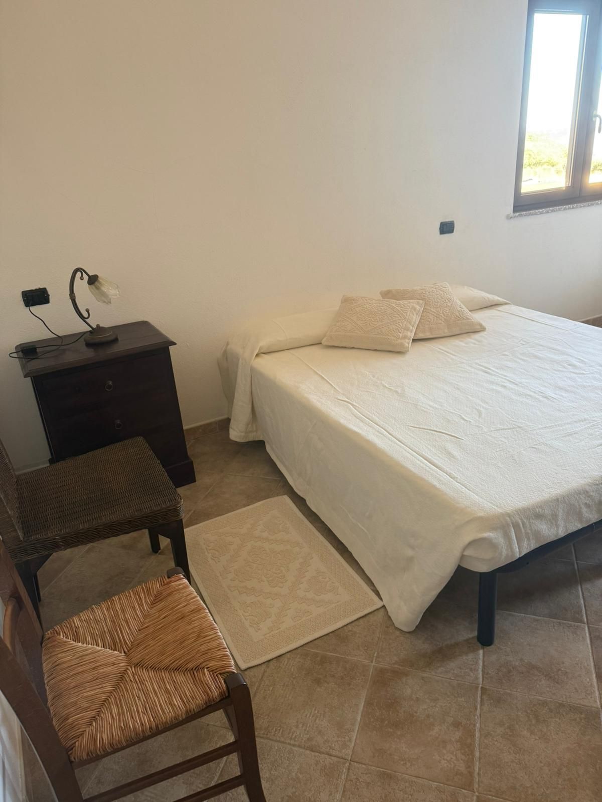 Piccola camera da letto con un letto bianco, due sedie, un comodino scuro e una finestra sulla destra.