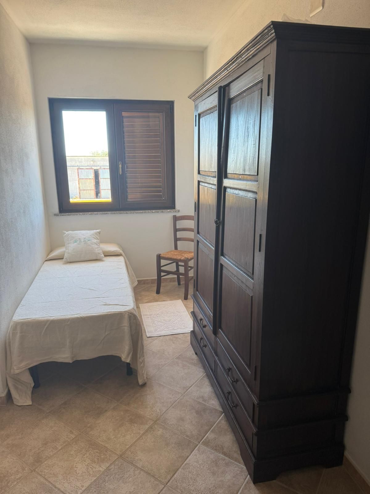 Piccola camera da letto con letto singolo, finestra, sedia e grande armadio scuro su pavimento piastrellato.
