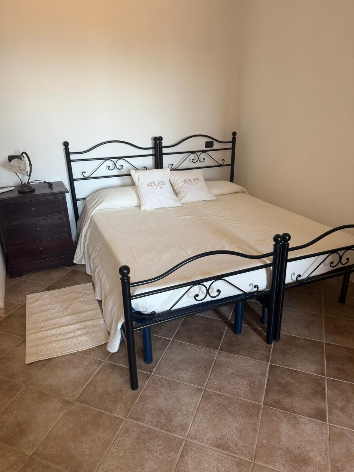 Camera da letto con letto in metallo nero, biancheria da letto beige, comodino e pavimento piastrellato.