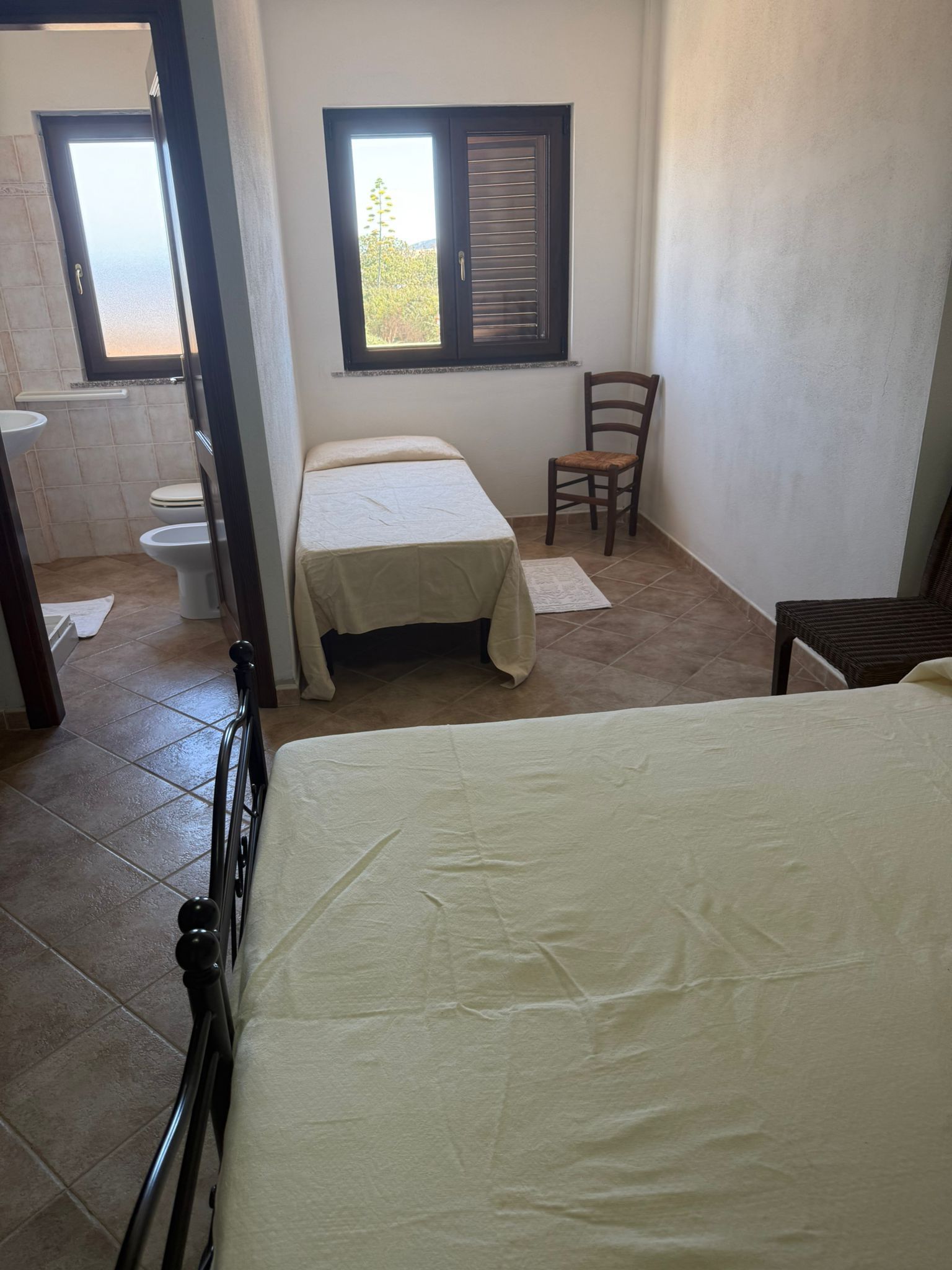 Piccola camera da letto con due letti singoli, sedia, finestra e porta del bagno aperta.