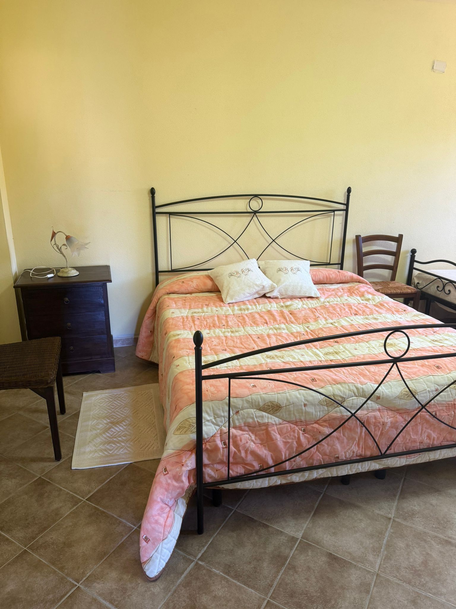 Camera da letto con letto in metallo nero, trapunta color pesca, cuscini, comodino, sedia e pavimento piastrellato su pareti gialle.