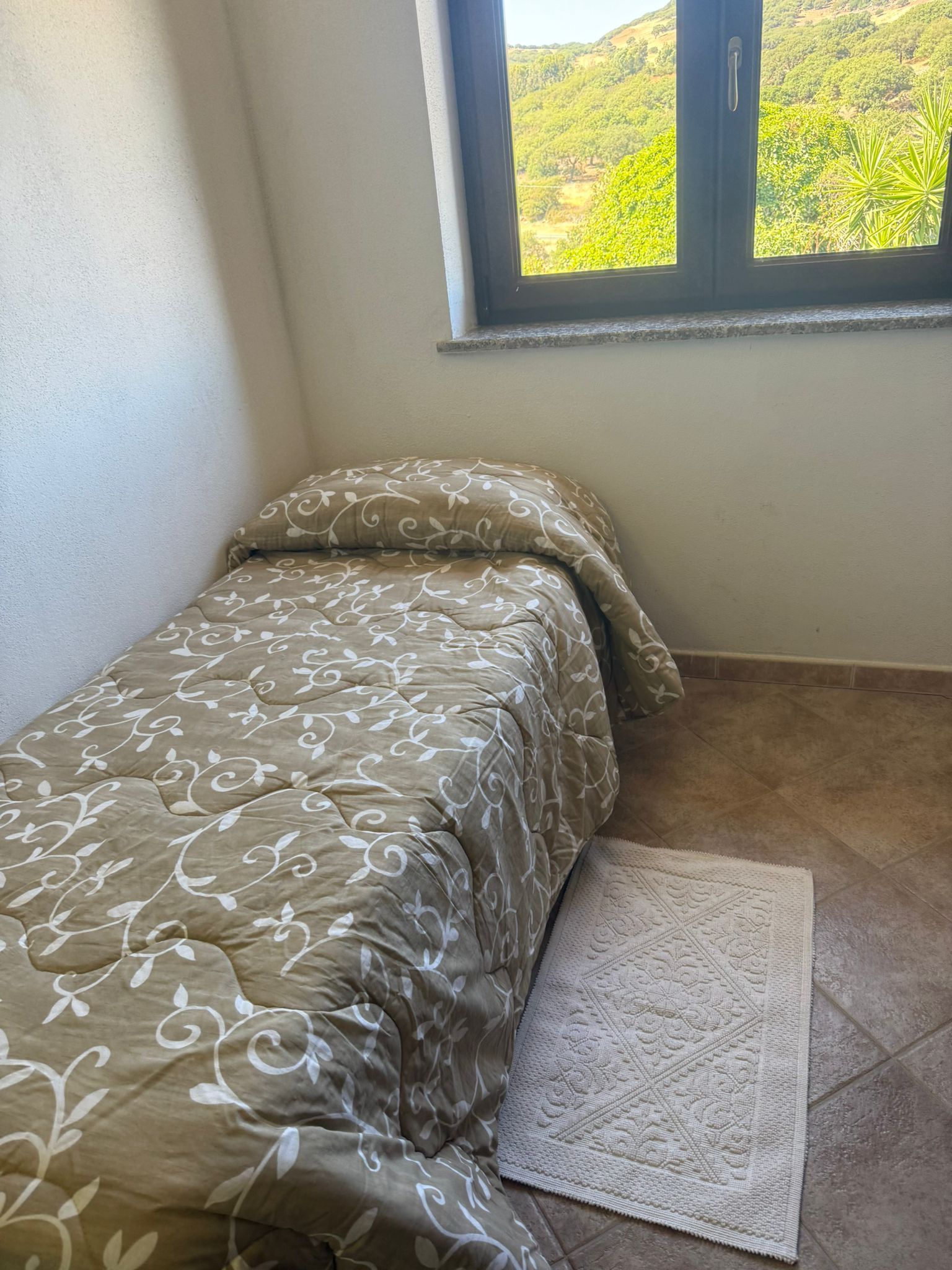 Piccola camera da letto con un letto singolo, lenzuola grigie a fantasia, un tappeto e una finestra che lascia entrare la luce del sole.