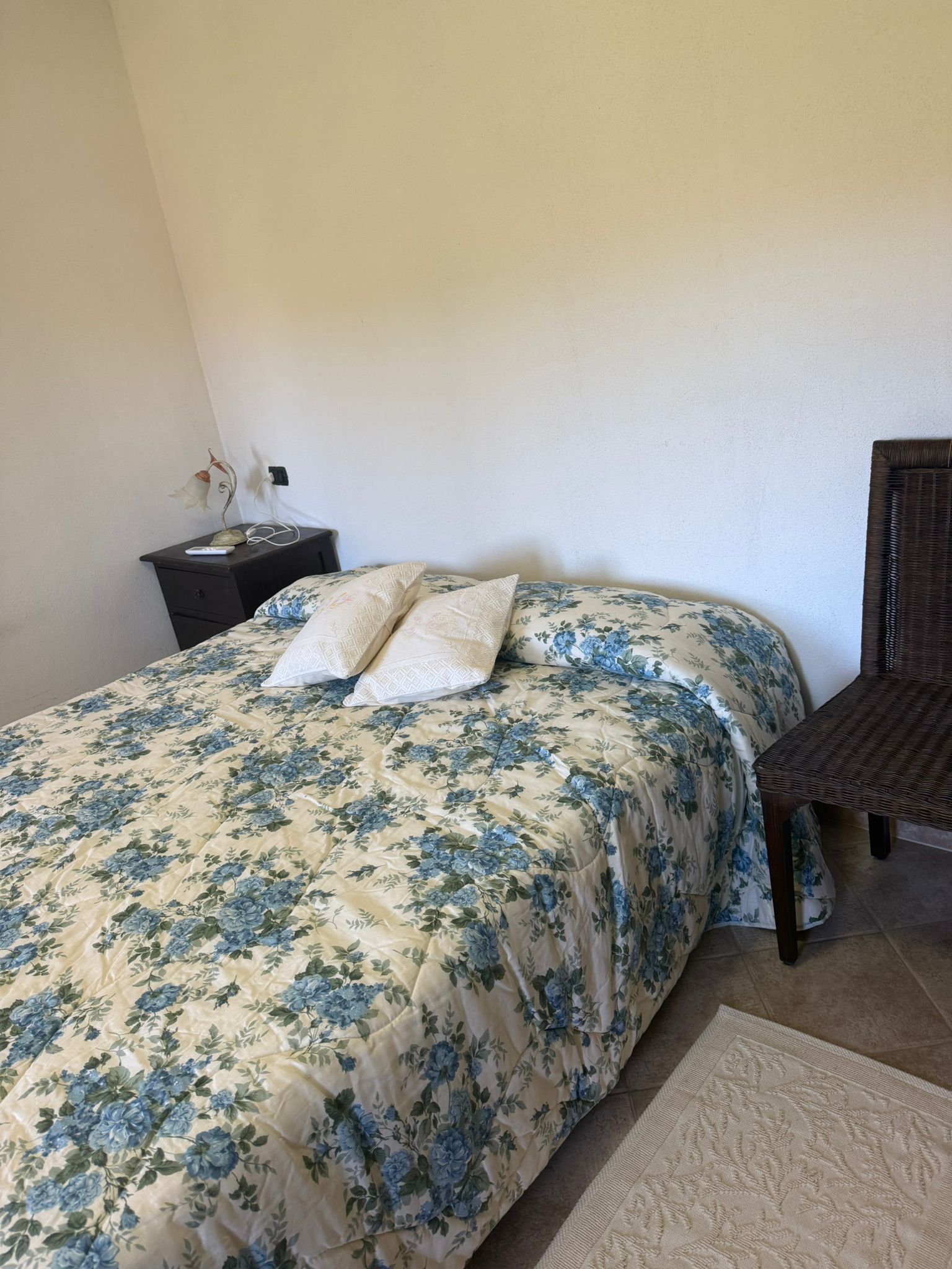 Camera da letto con copriletto blu a fiori, cuscini, comodino e sedia intrecciata su un tappeto.