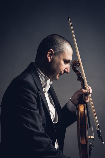 A musician in a tuxedo poses in profile against a dark background, holding a violin and bow near their face.