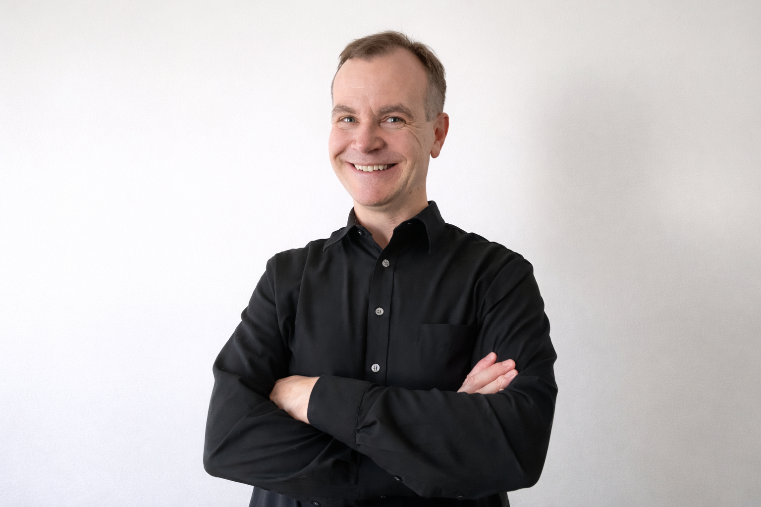 Zachary Stoughton in a black shirt standing against a white background with their arms crossed, smiling.