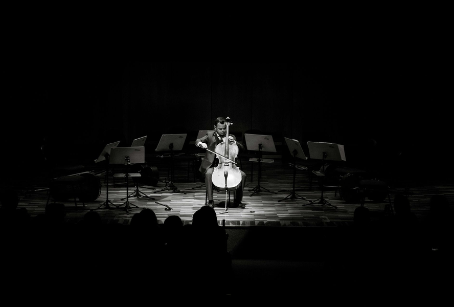 A spotlight shines on a cellist performing solo on a dark stage with music stands surrounding them.