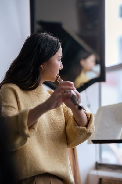 A person wearing a yellow sweater plays the flute while looking at music on a stand, reflected in a nearby mirror.
