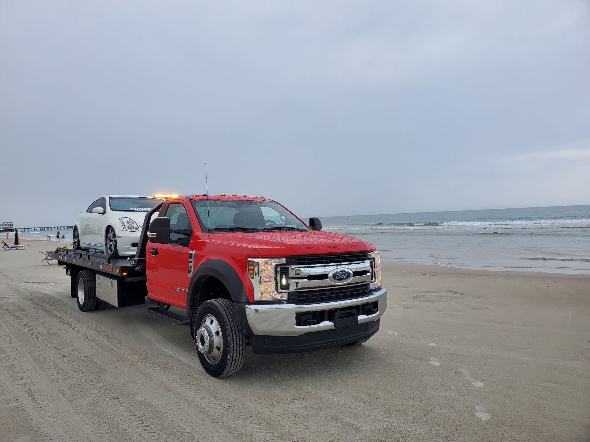 A red tow truck is towing a white car on a beach.