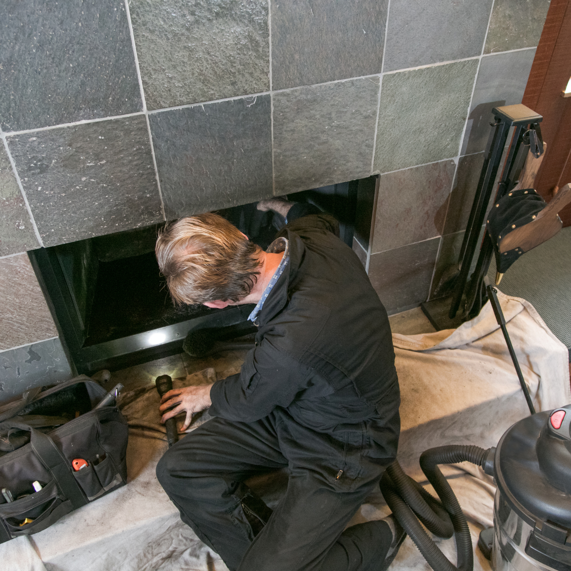 Person in black suit cleaning a fireplace, looking inside. Tools, black bag, and vacuum nearby.