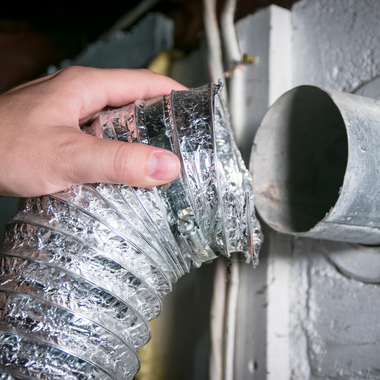 Hand connecting a flexible silver dryer vent hose to a metal vent in a white-painted room.