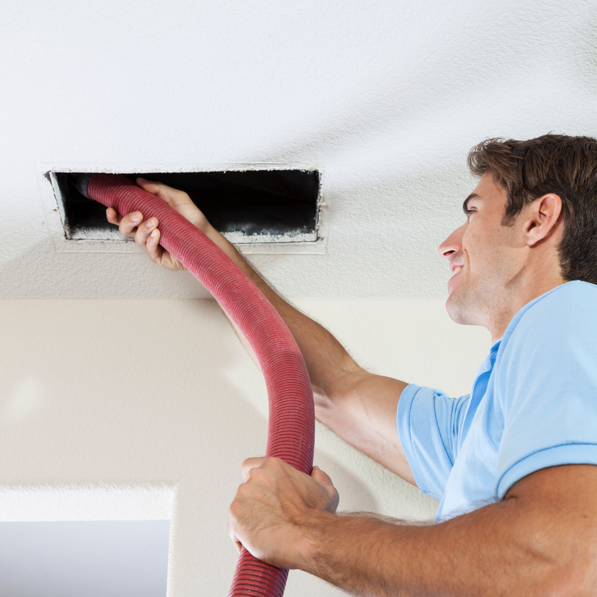 Man cleaning air vent in a white room using a red hose, smiling.