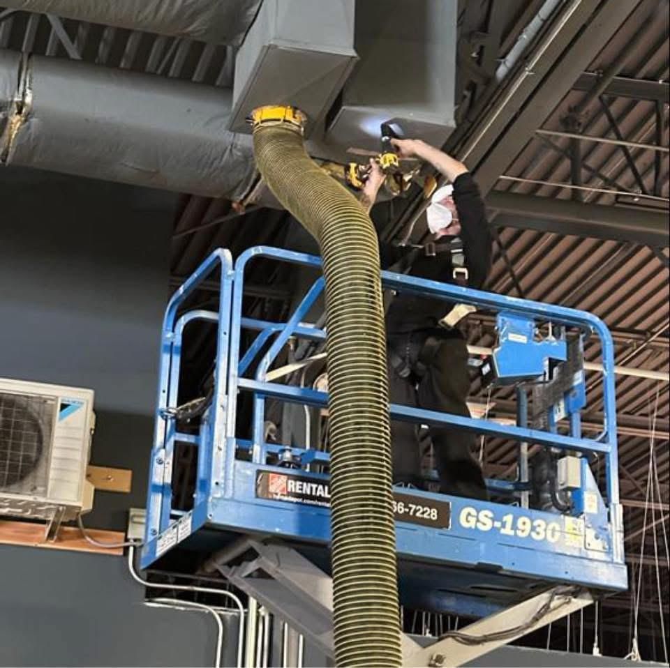 Person in lift cleaning ductwork with a hose and flashlight; industrial setting with exposed pipes and AC unit.