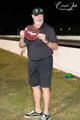A person wearing a black shirt, shorts, and a cap stands at a race track holding a round red award at night.