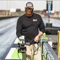 A person in a black shirt and cap holds a camera on a tripod at a drag racing track.