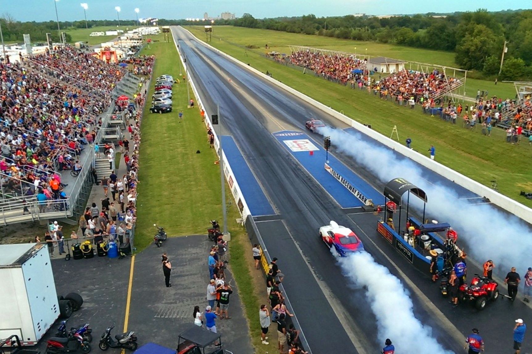 An aerial view of a drag strip race with cars performing burnouts, surrounded by a large crowd in outdoor bleachers.