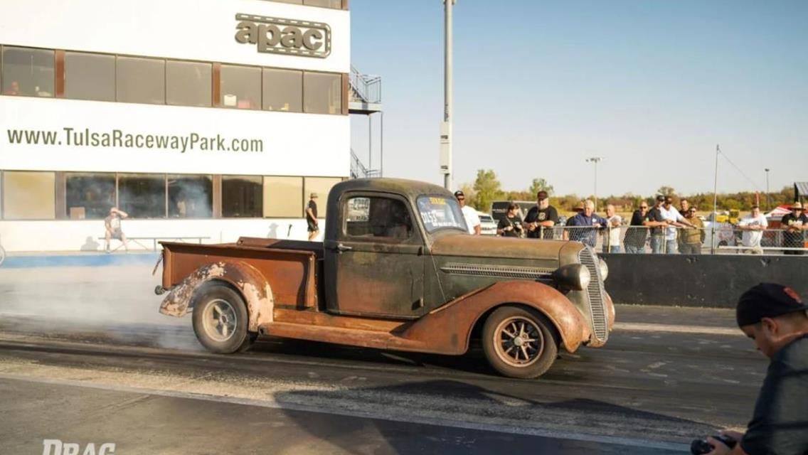 A rusted vintage pickup truck performing a burnout on a drag strip at Tulsa Raceway Park with spectators in the background.