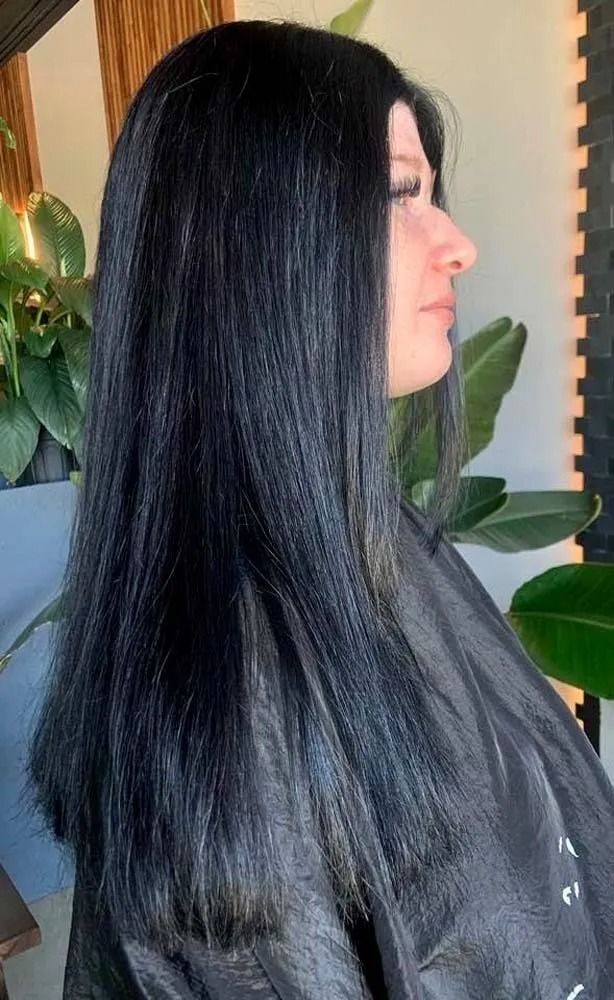 Profile view of a person with long black hair indoors, near green plants and a window