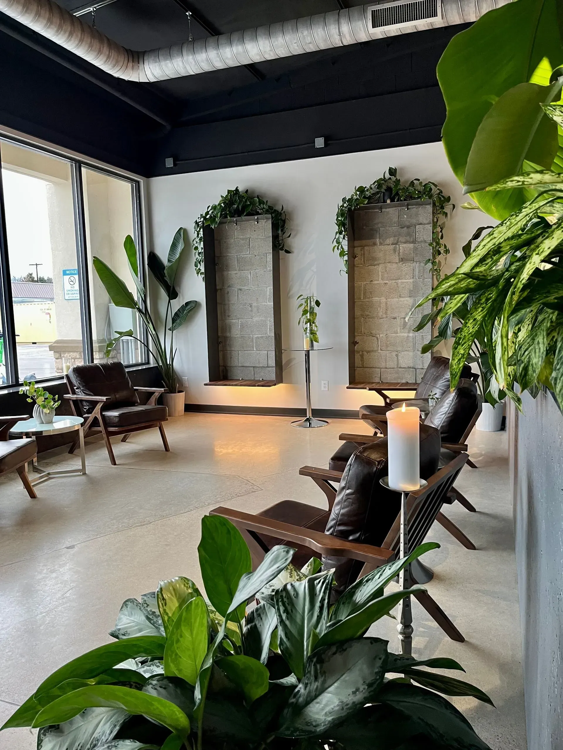 Modern lounge with wooden chairs, tall candles, lush plants, and vine-covered wall alcoves near large windows
