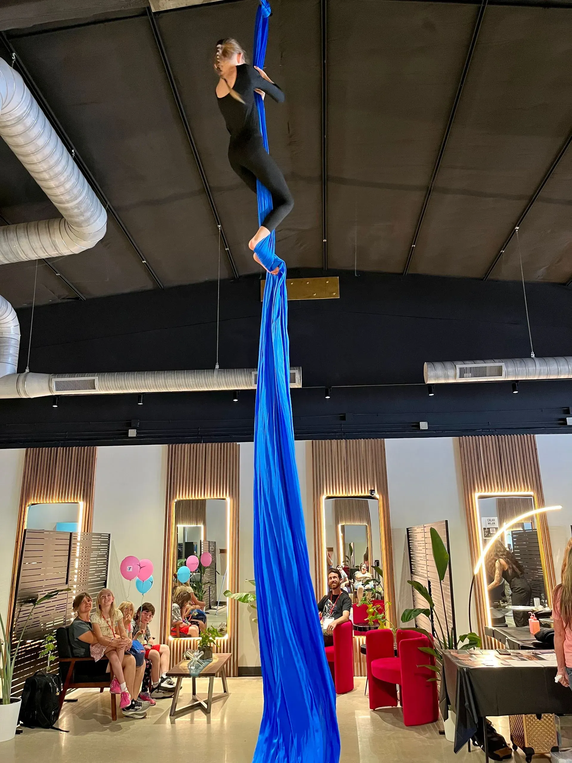 Aerial performer hanging in bright blue fabric inside a studio with spectators below.