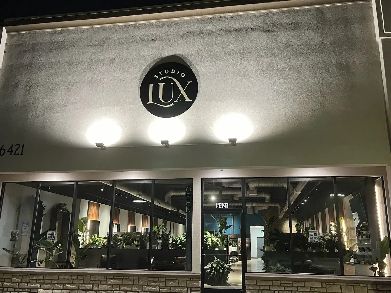 Night exterior of a modern storefront with a circular “JUX” sign and large glass windows lit inside.