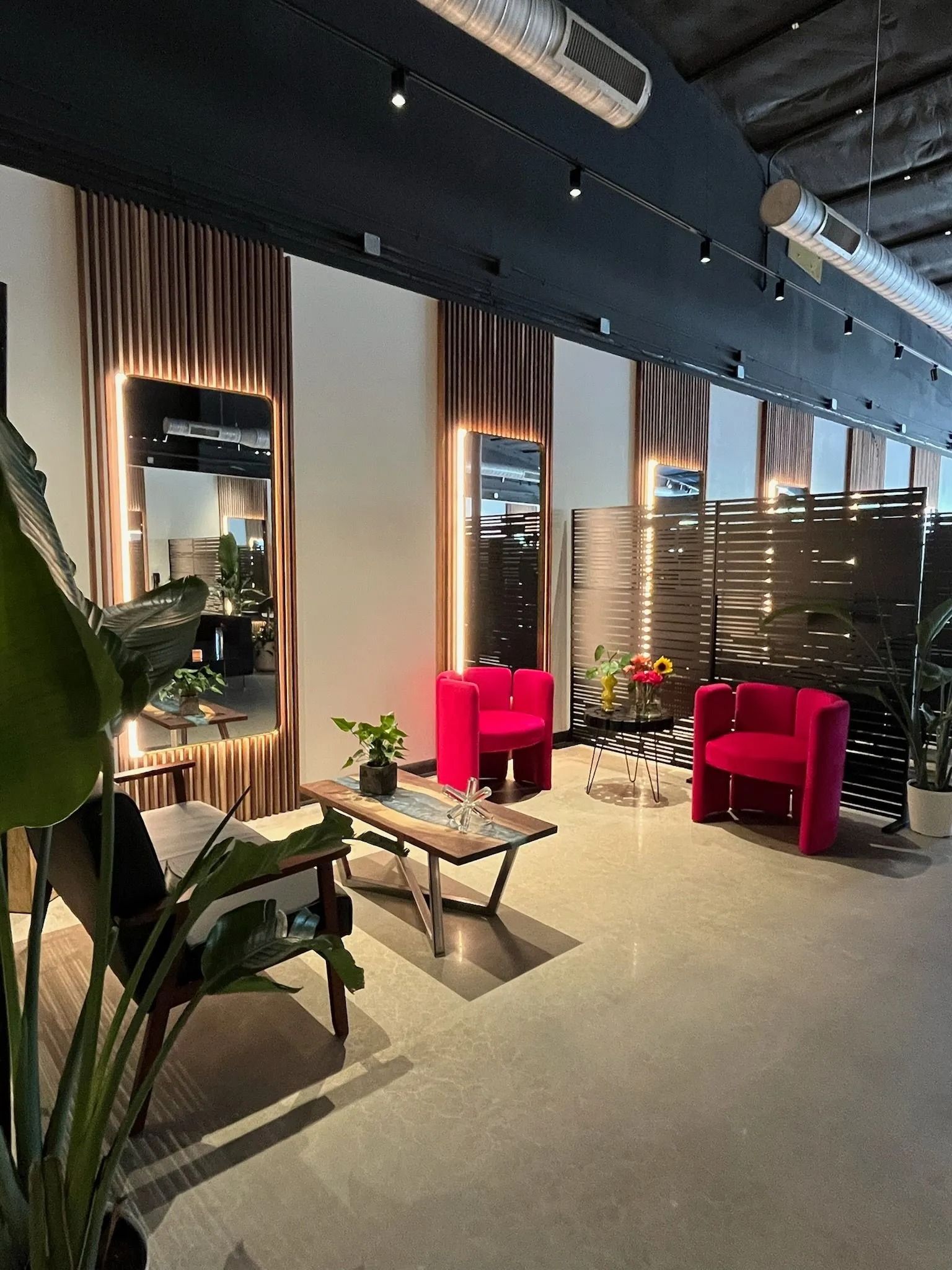 Modern lounge with pink chairs, wooden tables, mirrors, and tall curtains under a dark ceiling.