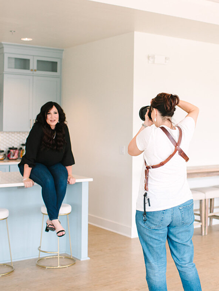 Woman poses on a light blue kitchen island while a photographer takes a photo; white room with stools.