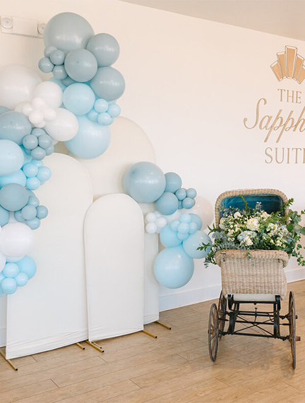 Baby shower decor with blue and white balloons, arch backdrop, and vintage stroller filled with flowers.