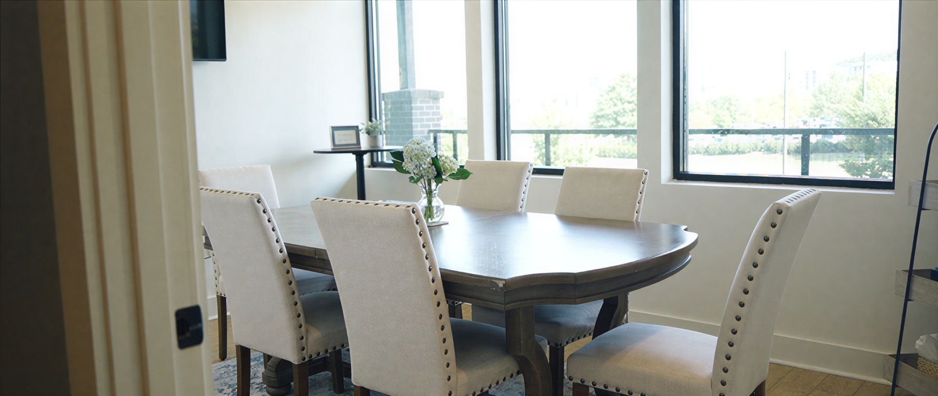 Dining room with oval table, six upholstered chairs, and large windows with outdoor view.