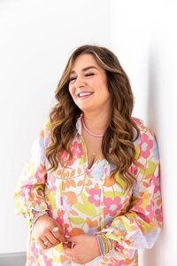 Woman smiling, wearing a floral-patterned shirt, leaning against a white wall.