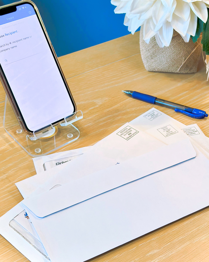 Phone on stand, envelopes, pen on a wooden desk with a white flower in the background.