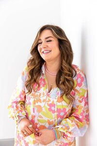 Woman leaning against a white wall, smiling, wearing a floral shirt and pink necklace.