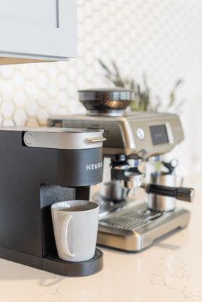 Black Keurig coffee maker with a gray mug next to a silver espresso machine on a countertop.