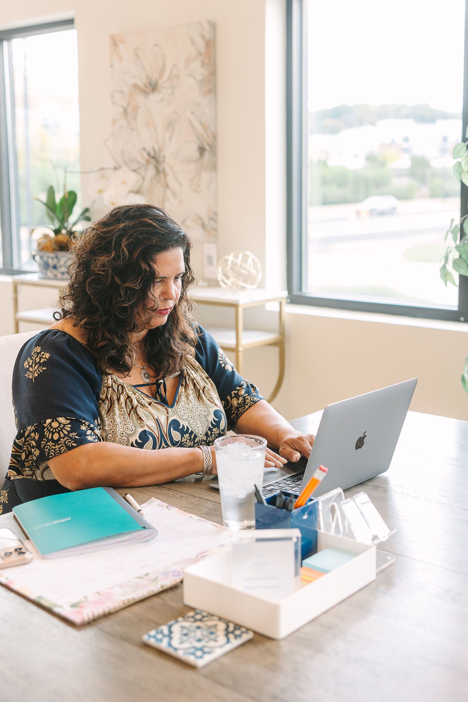 Woman at a desk with a laptop, working. There's a glass of water, notepad, and other office supplies. Bright room with a window.
