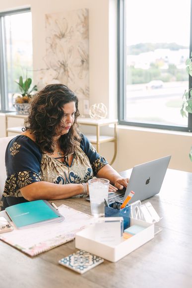 Woman at a desk with a laptop, working. There's a glass of water, notepad, and other office supplies. Bright room with a window.