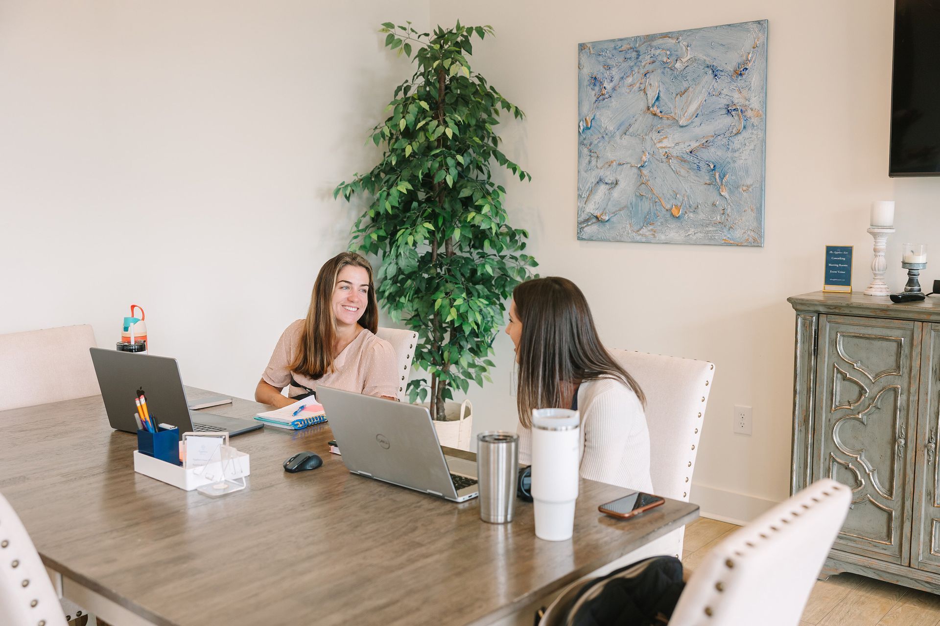 Two women sit at a table with laptops, talking. One smiles; the other looks on. There's a plant and art in the room.