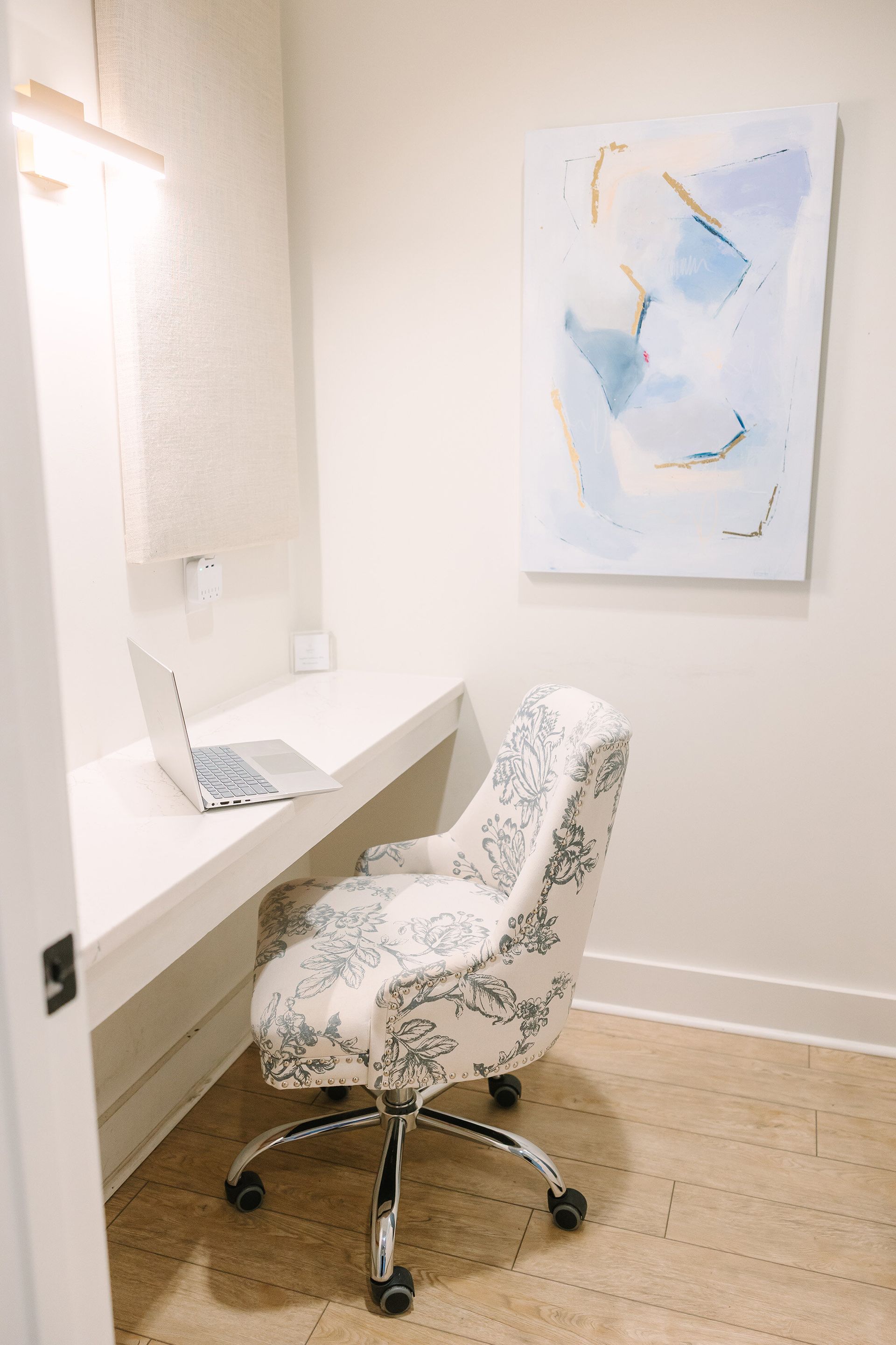 Desk with laptop, floral patterned chair on wheels, in a small office nook. Light wood floor, white desk.