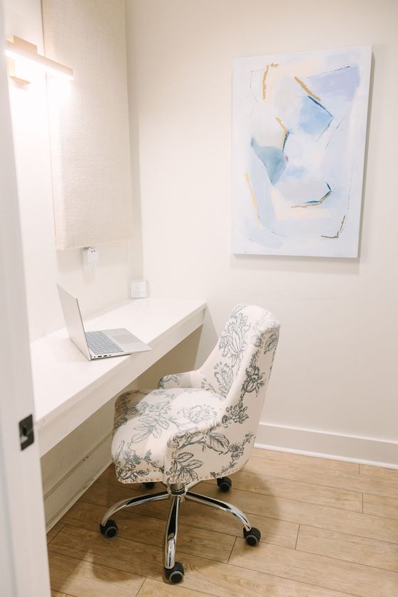 Desk with laptop, floral patterned chair on wheels, in a small office nook. Light wood floor, white desk.