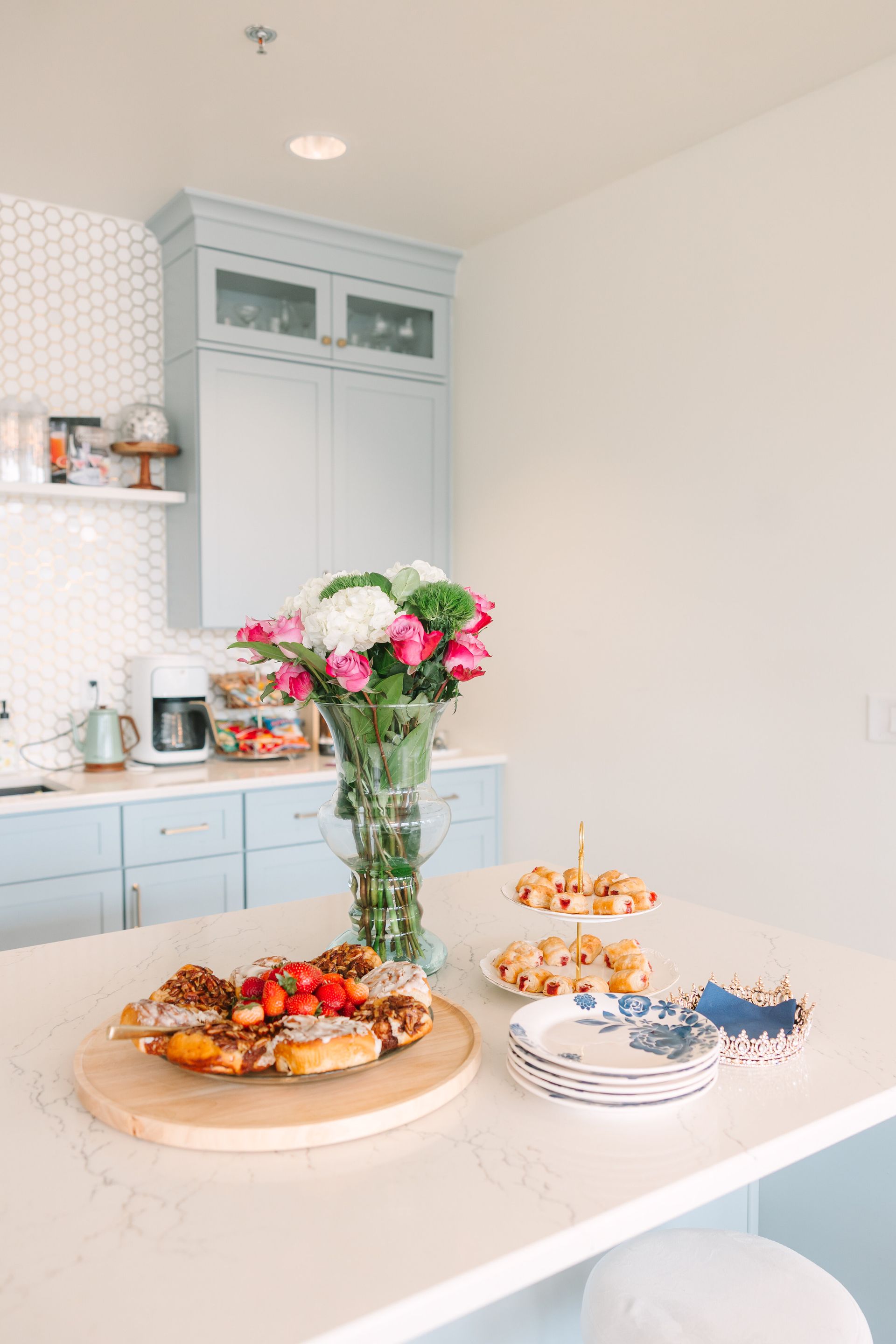 Kitchen island with flowers, pastries, and appetizers, near light blue cabinets and white walls.