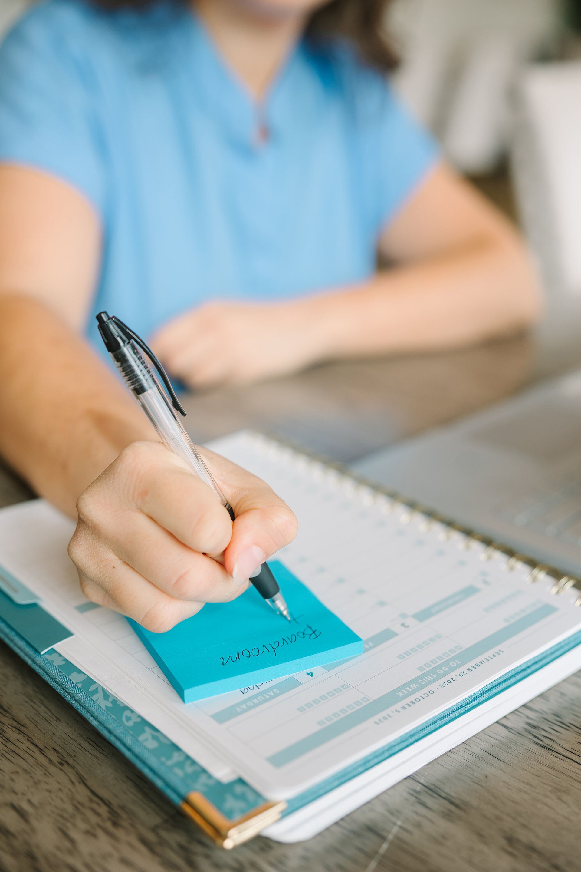 Person writing with a pen in a notebook with a blue sticky note attached on a table.