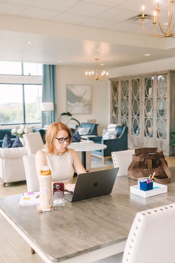 Woman working on a laptop at a large table in a bright, elegant room.